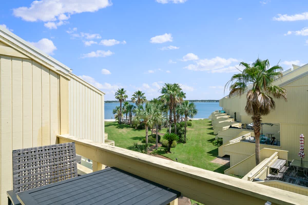 View from a balcony showing a dark grey outdoor table and chair, overlooking a lush green lawn with palm trees, leading to a calm blue body of water under a partly cloudy sky. Yellow-sided townhouses line the sides of the view.