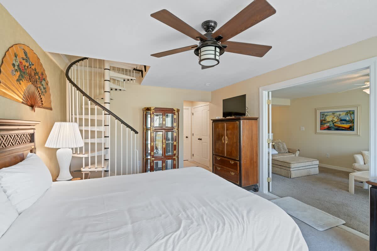 Bedroom with a queen bed, dark wood furniture, and a spiral staircase, with French doors opening to the living room and a TV on a cabinet.