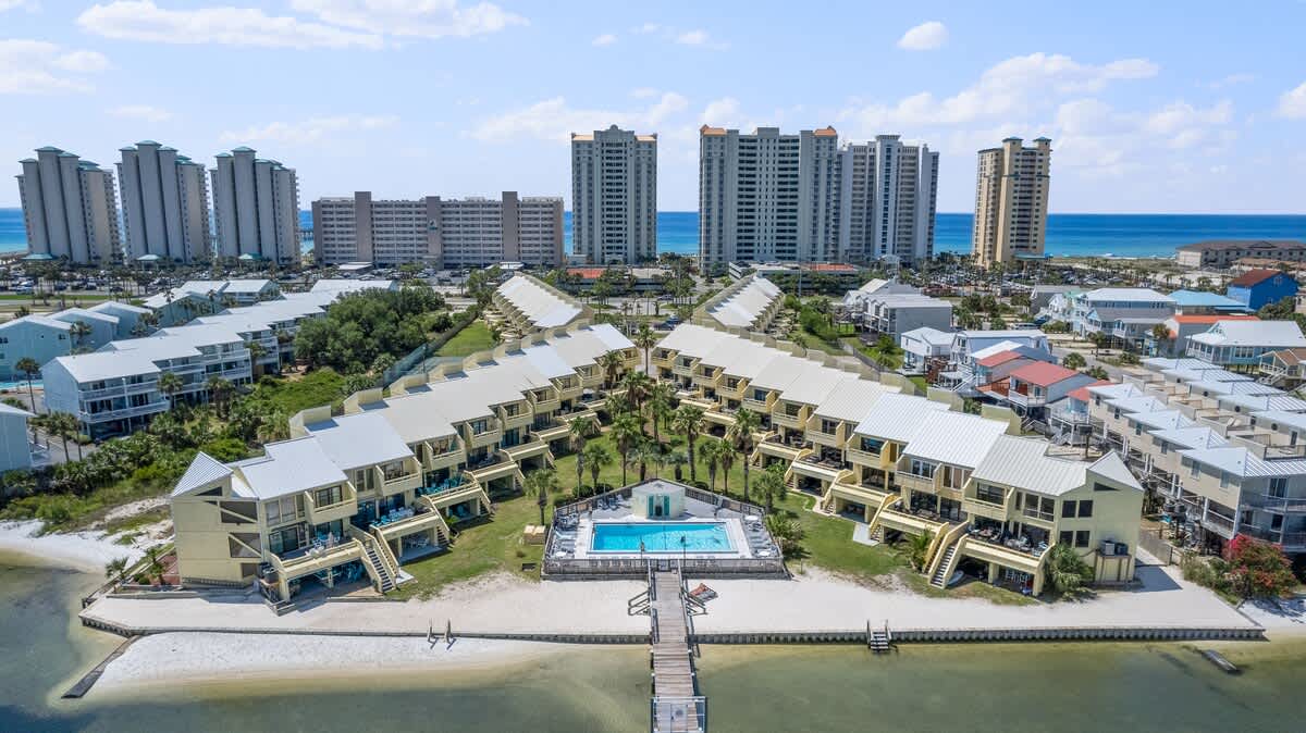 Closer aerial view of the townhouse complex, focusing on the rectangular swimming pool surrounded by a patio, and a long wooden dock with boat slips. A small sandy beach area is visible along the waterfront, with high-rise buildings in the background.