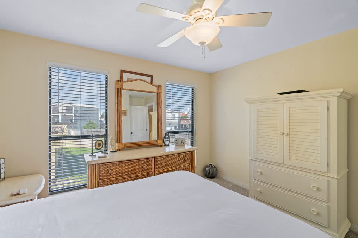 This view of Bedroom 1 highlights the queen bed, a large dresser with a mirror, and a white armoire, with multiple windows providing ample natural light and horizontal blinds for privacy.