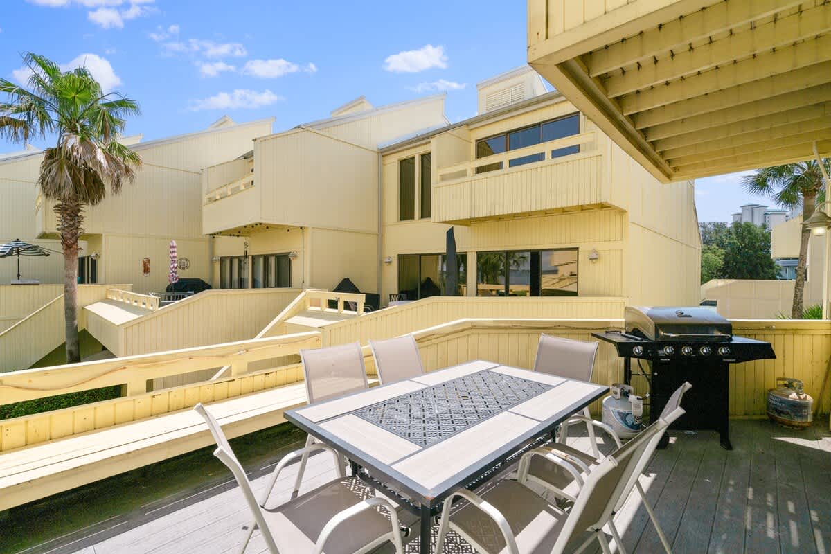 Outdoor deck with a large rectangular dining table and chairs, and a black gas BBQ grill. The deck is surrounded by yellow-sided townhouse structures, with a glimpse of palm trees and water in the background.