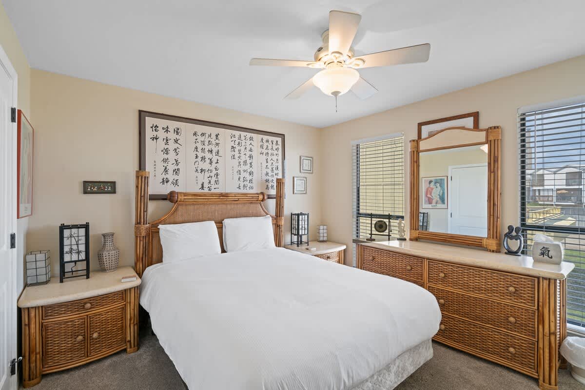 Bedroom 1 features a queen-sized bed with a rattan headboard, flanked by two matching nightstands, and a large dresser with a mirror, all set against light yellow walls with windows offering natural light.