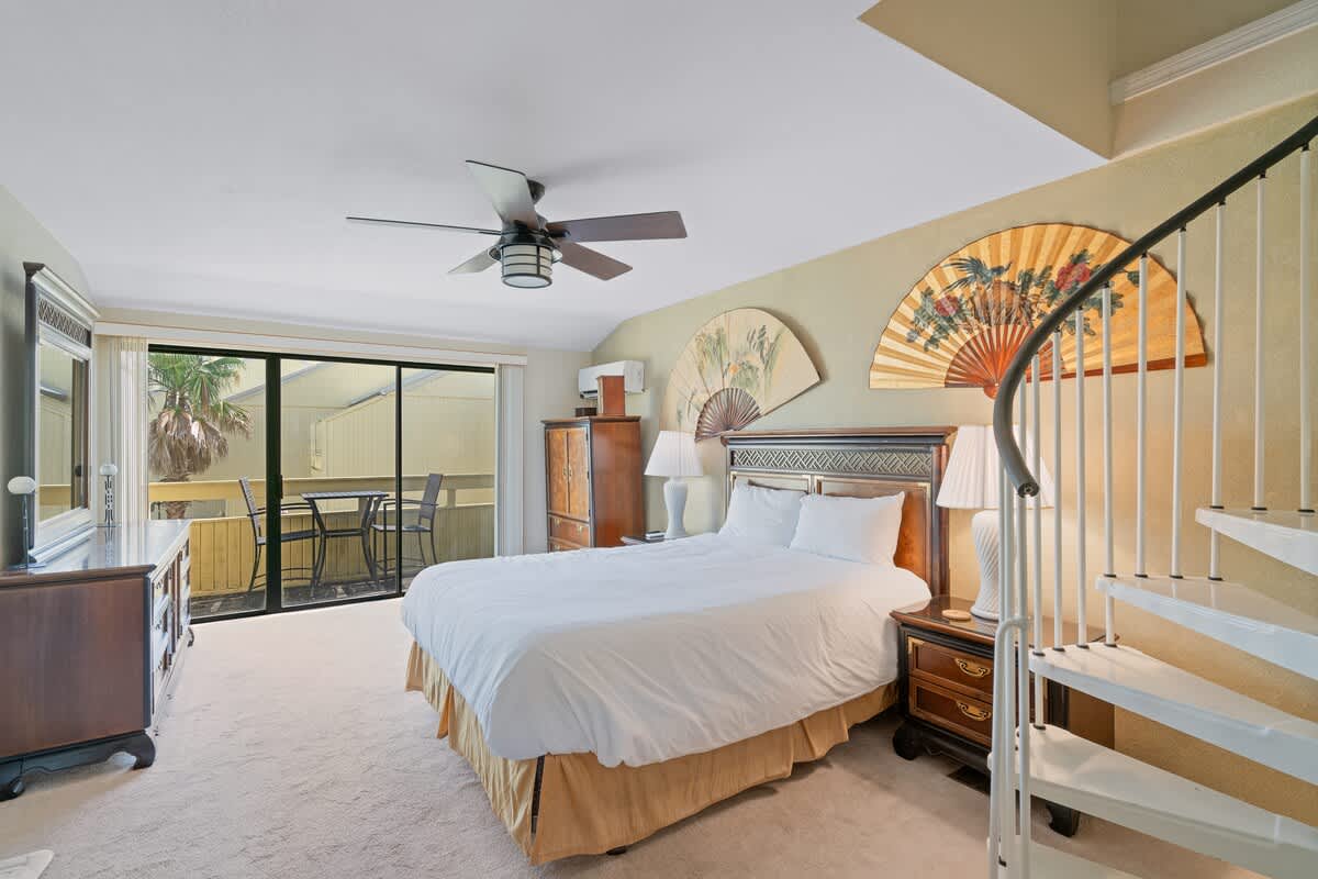 Bedroom with a queen bed, dark wood furniture, and a sliding glass door leading to a balcony with outdoor seating, featuring a spiral staircase.