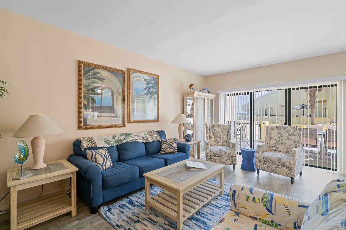 The bright living room features a blue sofa, two floral armchairs, a light wood coffee table, and a blue and white striped rug, with sliding glass doors leading to an outdoor area.