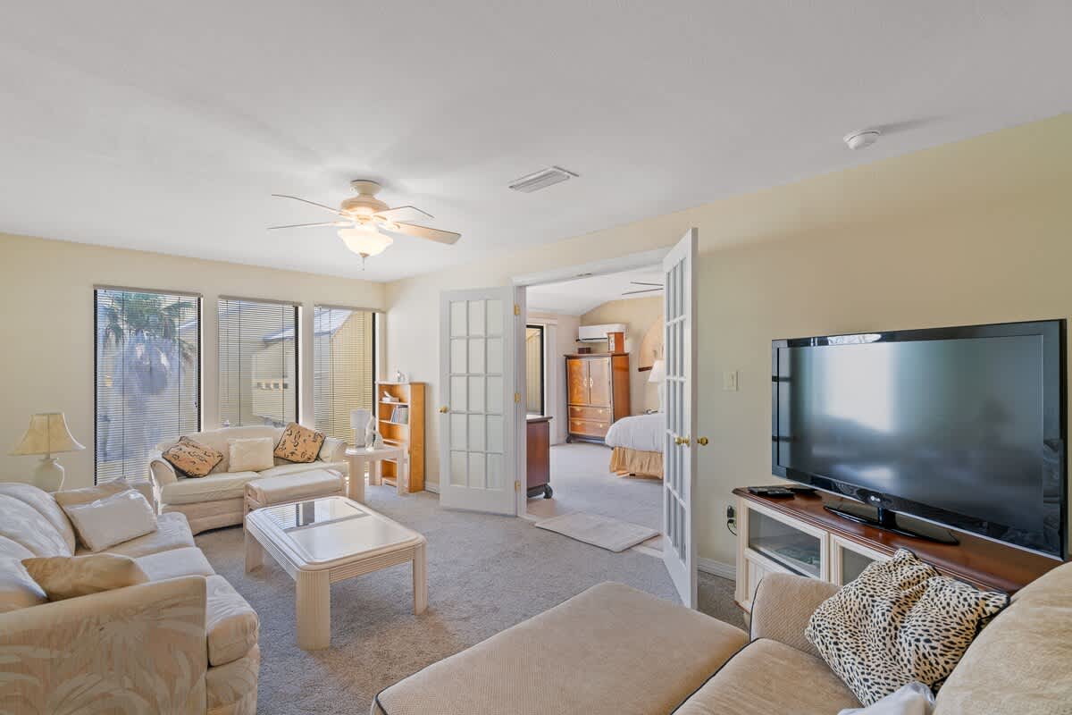 Cozy living room with light-colored sofas, a white coffee table, and a TV stand with a large flat-screen TV, connected to a bedroom via French doors.