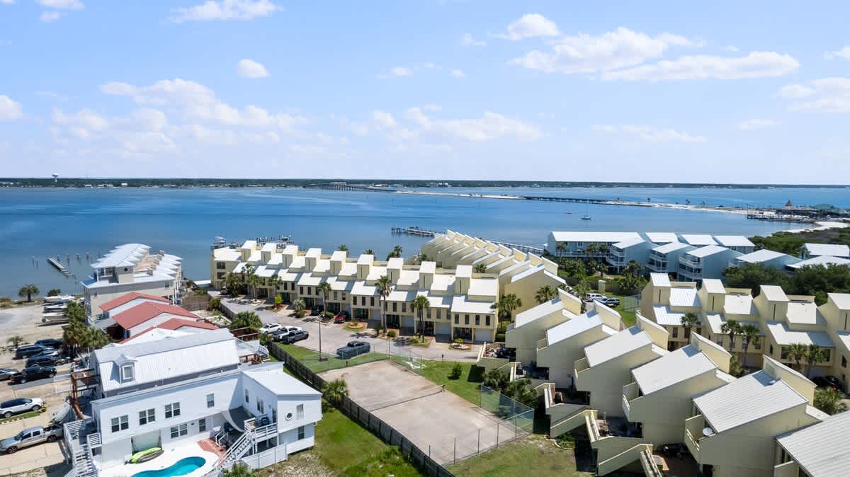 Broad aerial shot showcasing the townhouse complex, an adjacent tennis court, and the expansive bay with a distant bridge under a clear sky. Various residential buildings are scattered around.