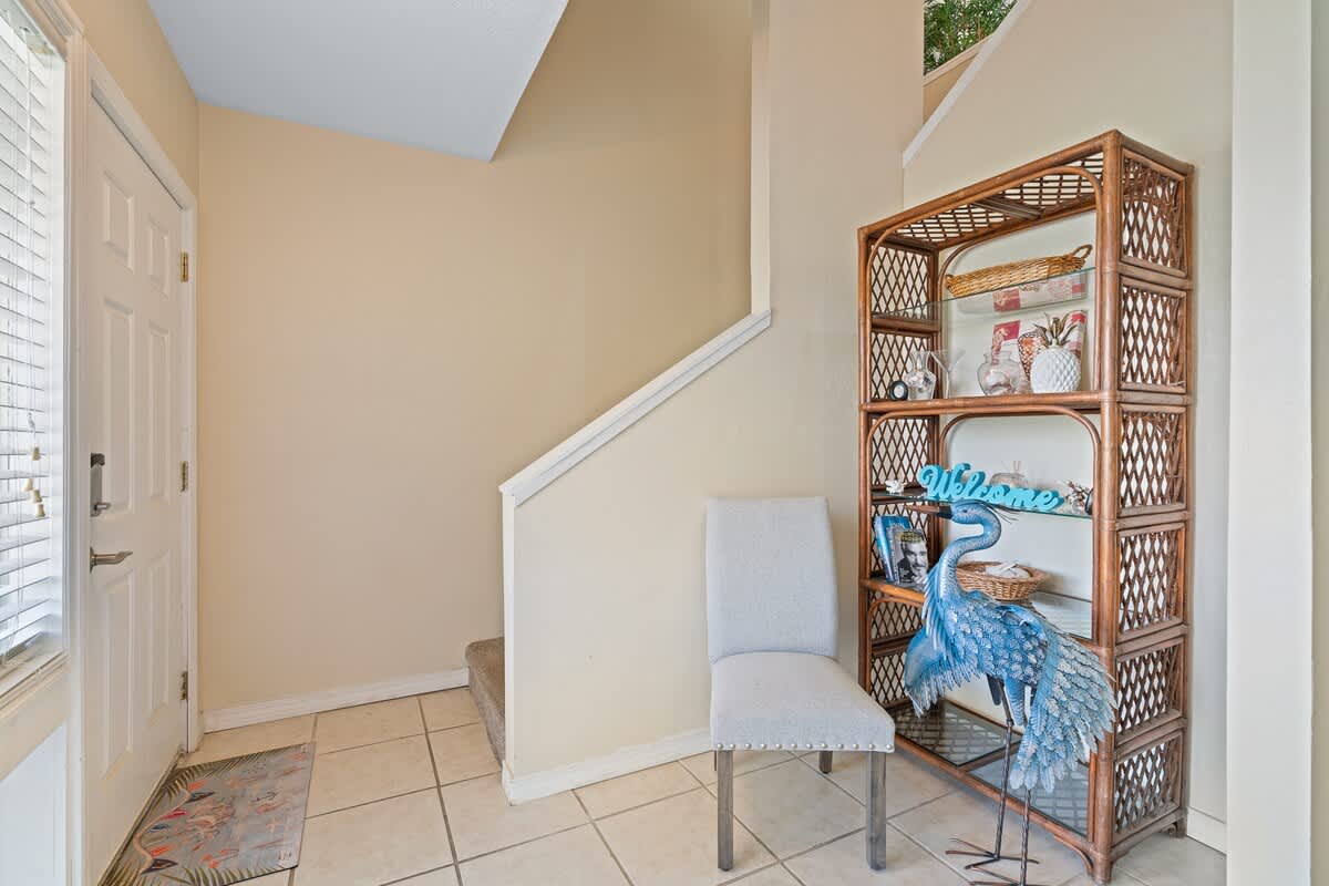 The bright foyer features light beige walls, white tiled flooring, a white front door with a window, and a wooden display shelf next to a light blue upholstered chair and a large blue heron statue.