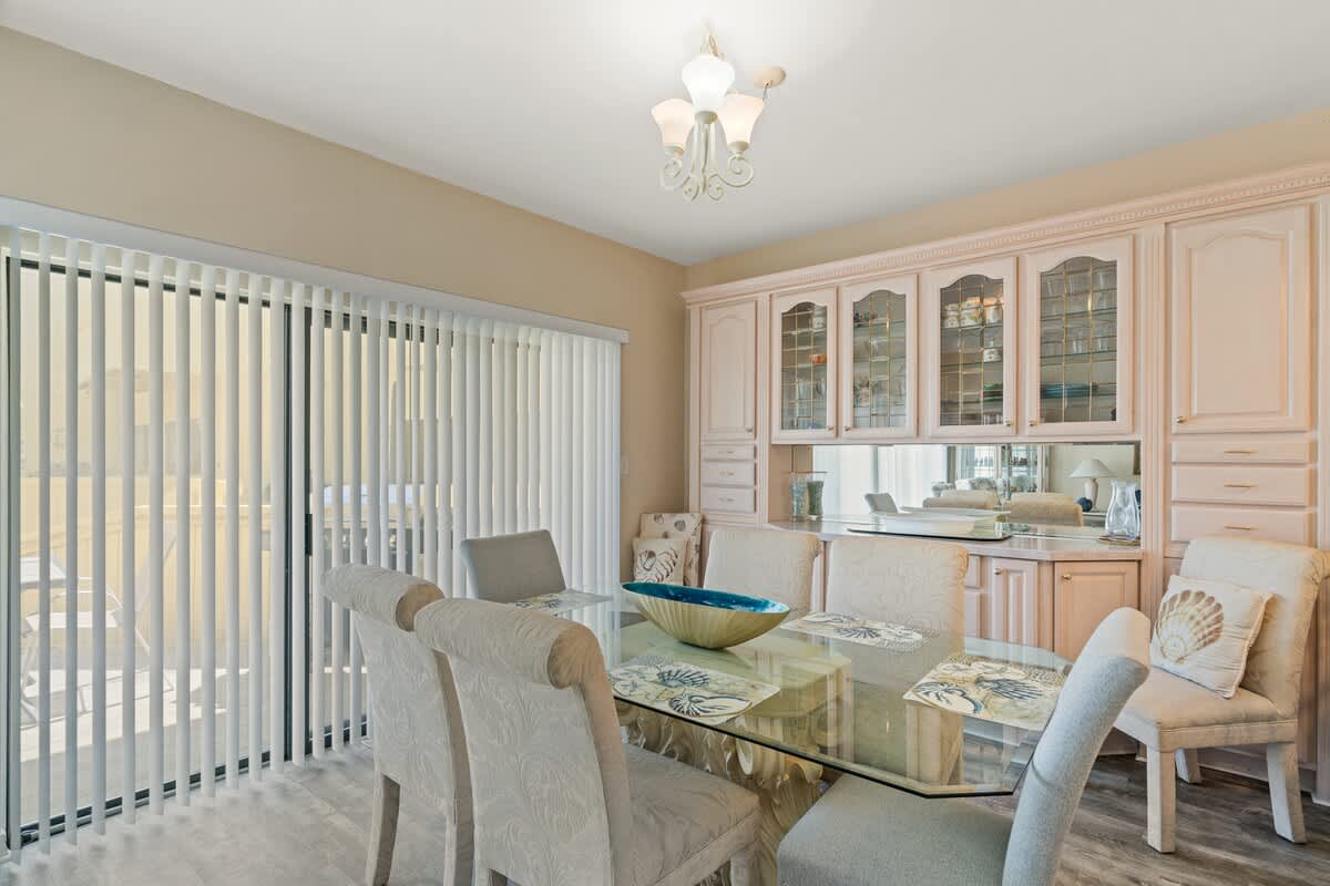A bright dining area features a glass-top table surrounded by six upholstered chairs, complemented by a light-colored built-in cabinet with glass doors and a large sliding glass door with vertical blinds.