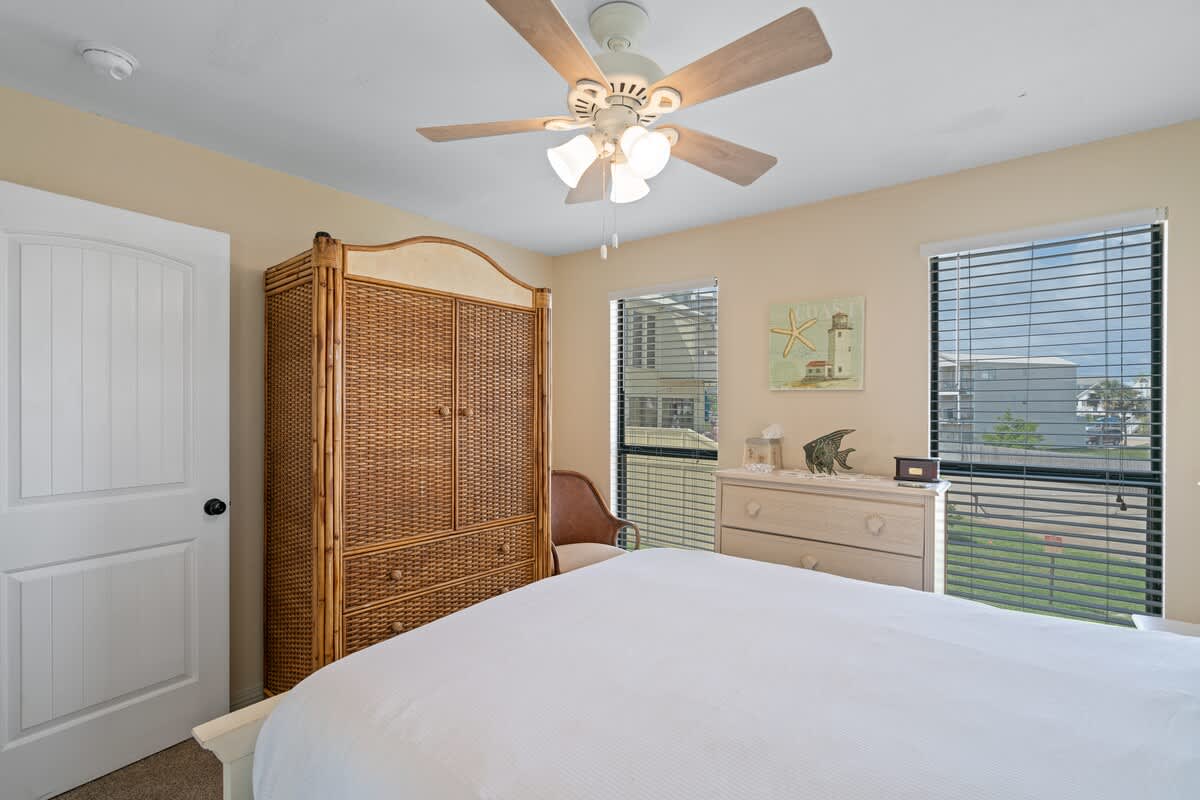 View of a bedroom with a large wicker armoire, a light-colored dresser with coastal decor, and two windows overlooking the exterior.