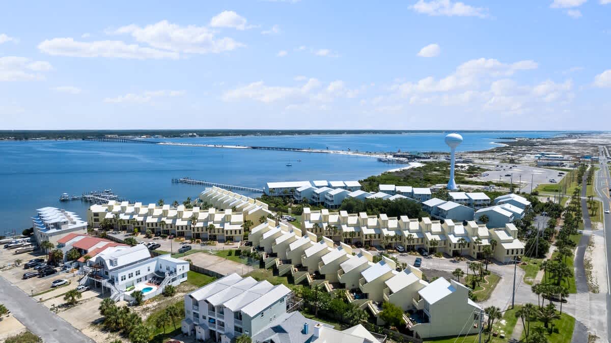 High aerial view showcasing a coastal town with a large body of water, a long bridge, and a water tower. Rows of light-colored townhouses are visible along the waterfront, with roads and other buildings in the foreground.