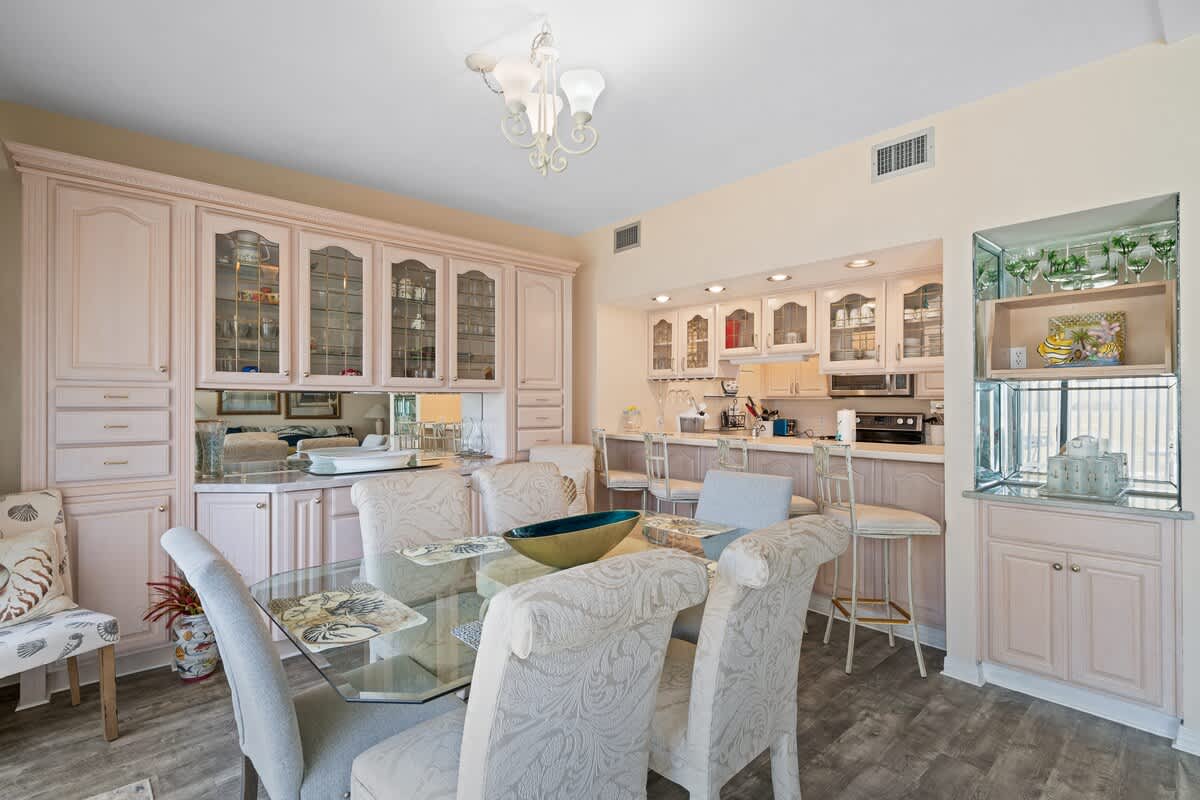 This dining space showcases a glass-top table with six upholstered chairs, positioned in front of an extensive light-colored built-in cabinet with mirrored and glass-front sections, adjacent to a kitchen bar area.