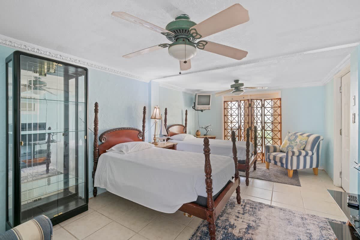 The bedroom features two dark wood four-poster twin beds, light blue walls, tiled flooring, a ceiling fan, and a tall glass display cabinet reflecting the room.