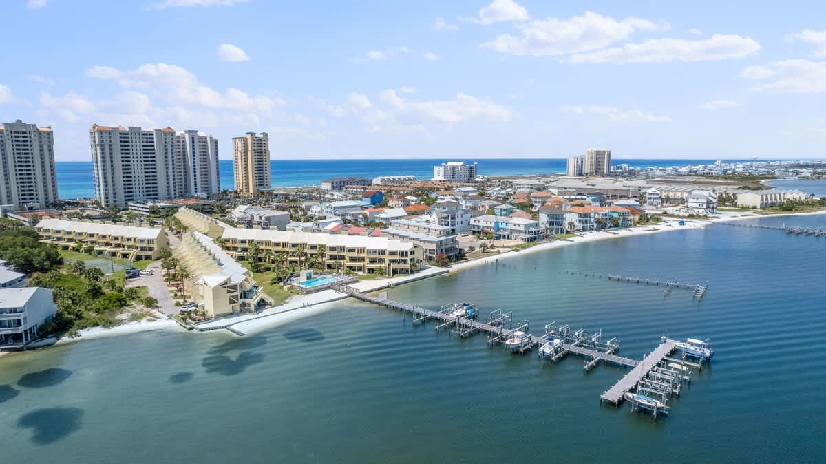 Panoramic aerial view of the coastal townhouse community, showing multiple yellow-sided buildings, a central swimming pool, and an extensive wooden dock with numerous boat lifts and slips. The calm bay water leads to the open ocean in the distance, with high-rise buildings along the coast.