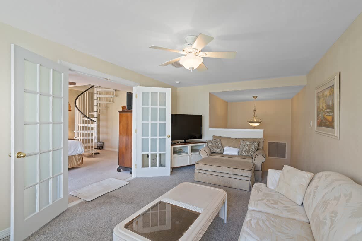 Living room with light-colored seating, a white coffee table, and a TV console, offering a glimpse into an adjacent bedroom with a spiral staircase.