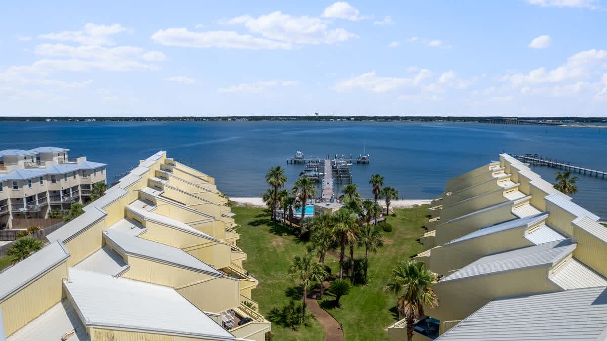 Aerial view looking down a lush green lawn flanked by two rows of yellow townhouses, leading directly to a private dock extending into the calm bay waters. Palm trees dot the landscape.