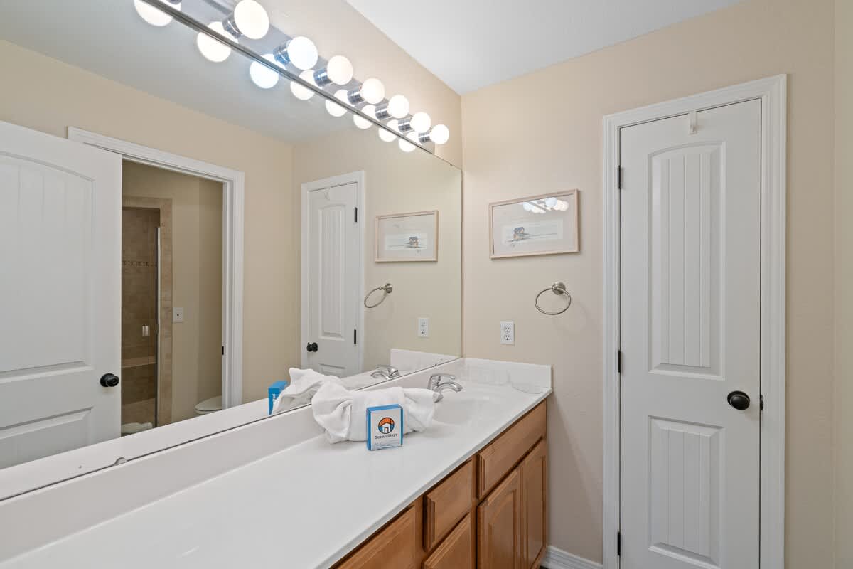 Bathroom vanity with a long white countertop, double sinks, a large mirror with Hollywood-style lights, and light wood cabinets.
