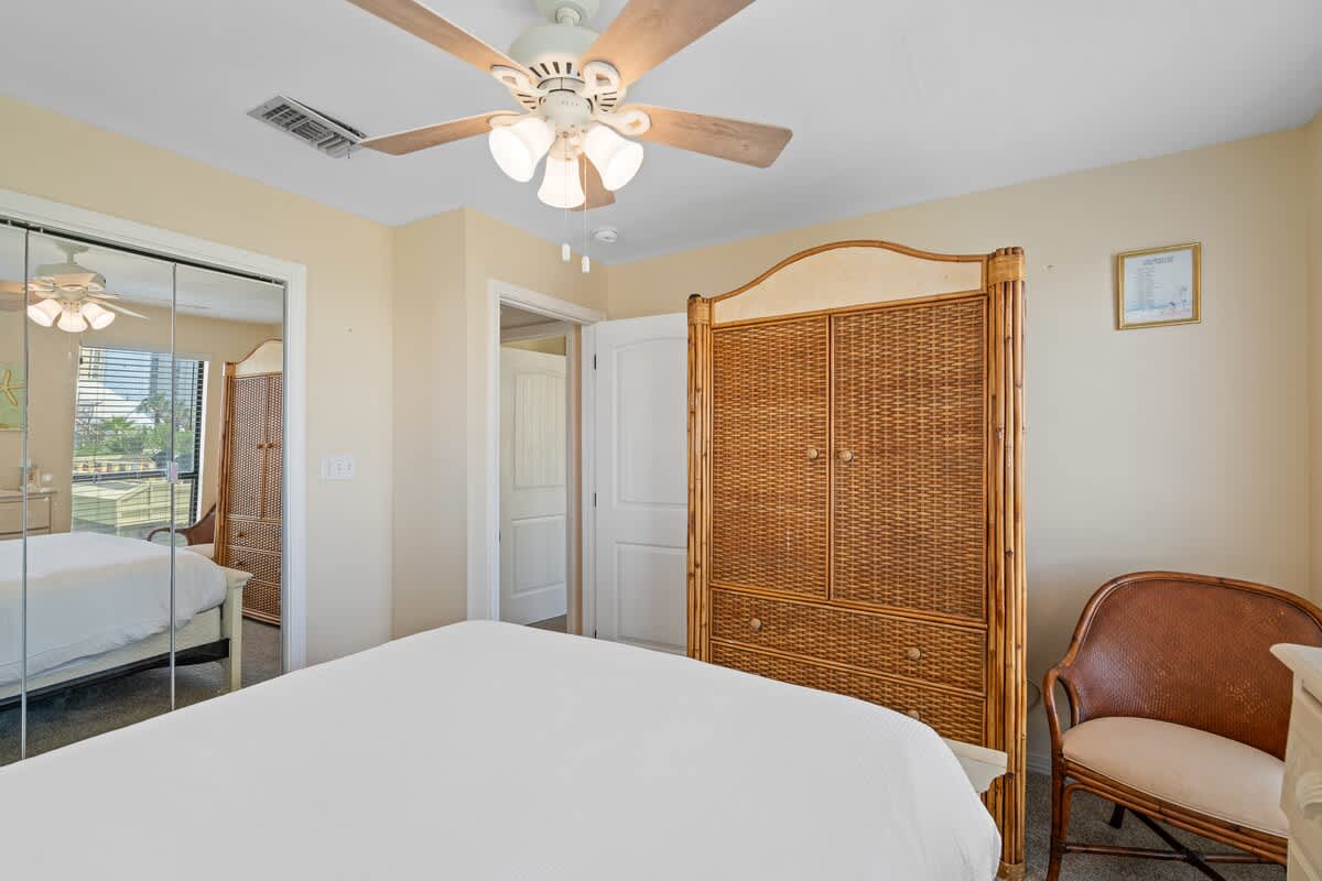 Bedroom view showing a mirrored closet reflecting the room, a wicker armoire, and a white door, with a ceiling fan overhead.