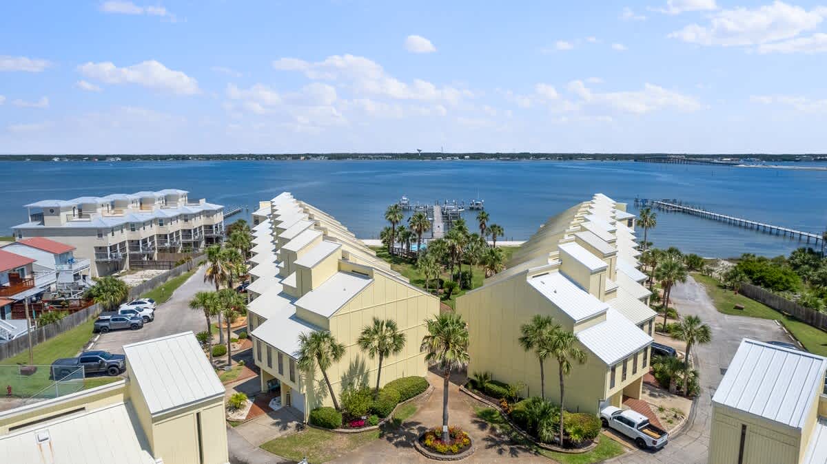 Aerial view of two rows of yellow-sided townhouses with white roofs, arranged around a central landscaped area with palm trees. A large body of water with a long pier is visible in the background.