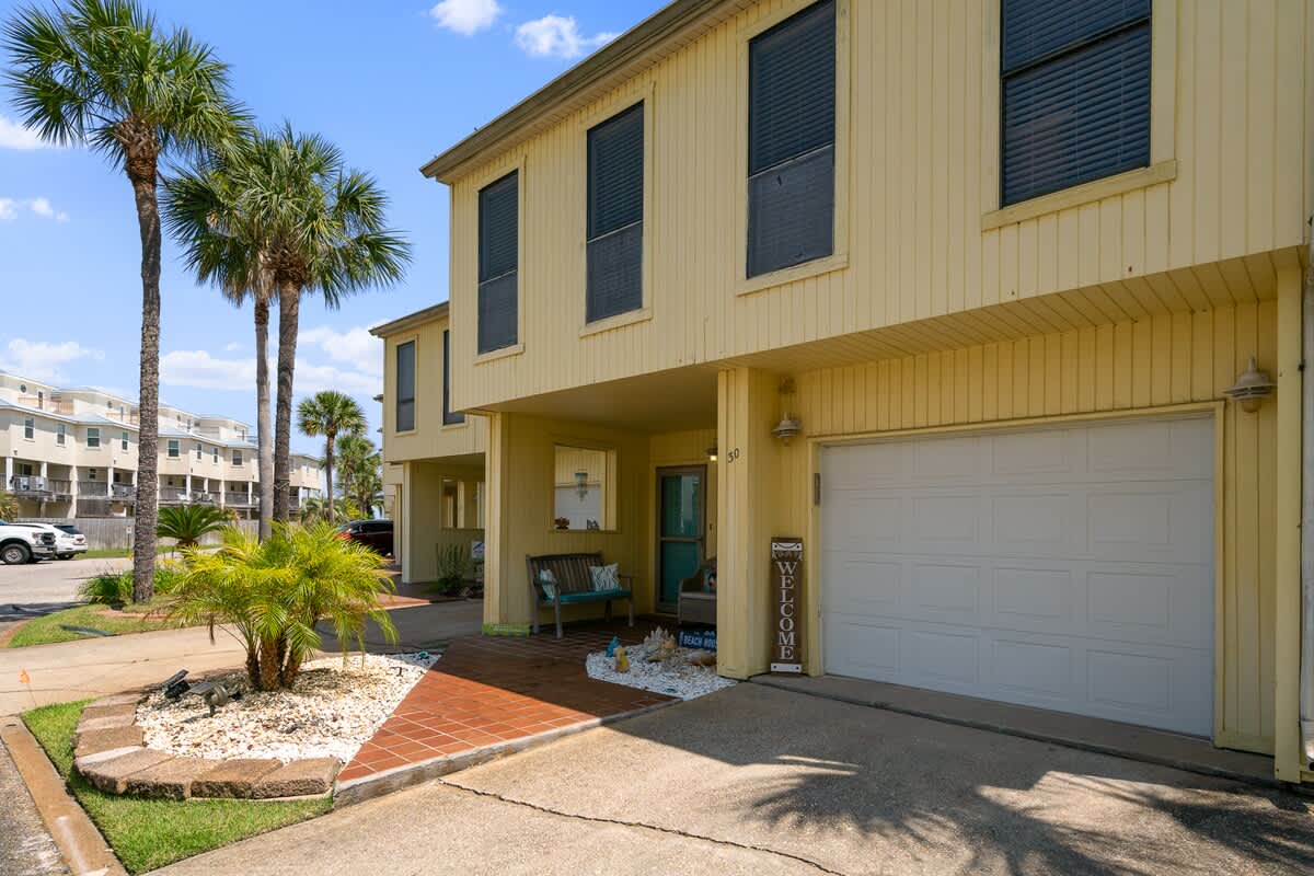 The front exterior of a two-story yellow townhouse features a white garage door, a covered porch entrance with a bench, and tall palm trees lining the driveway under a bright blue sky.