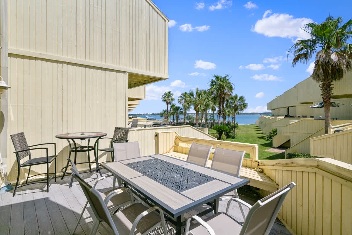 Spacious outdoor deck featuring a large rectangular dining table with six chairs, and a smaller round table with two chairs, all in dark grey and light grey tones. The deck overlooks a grassy area with palm trees and a body of water, with yellow-sided townhouses visible.