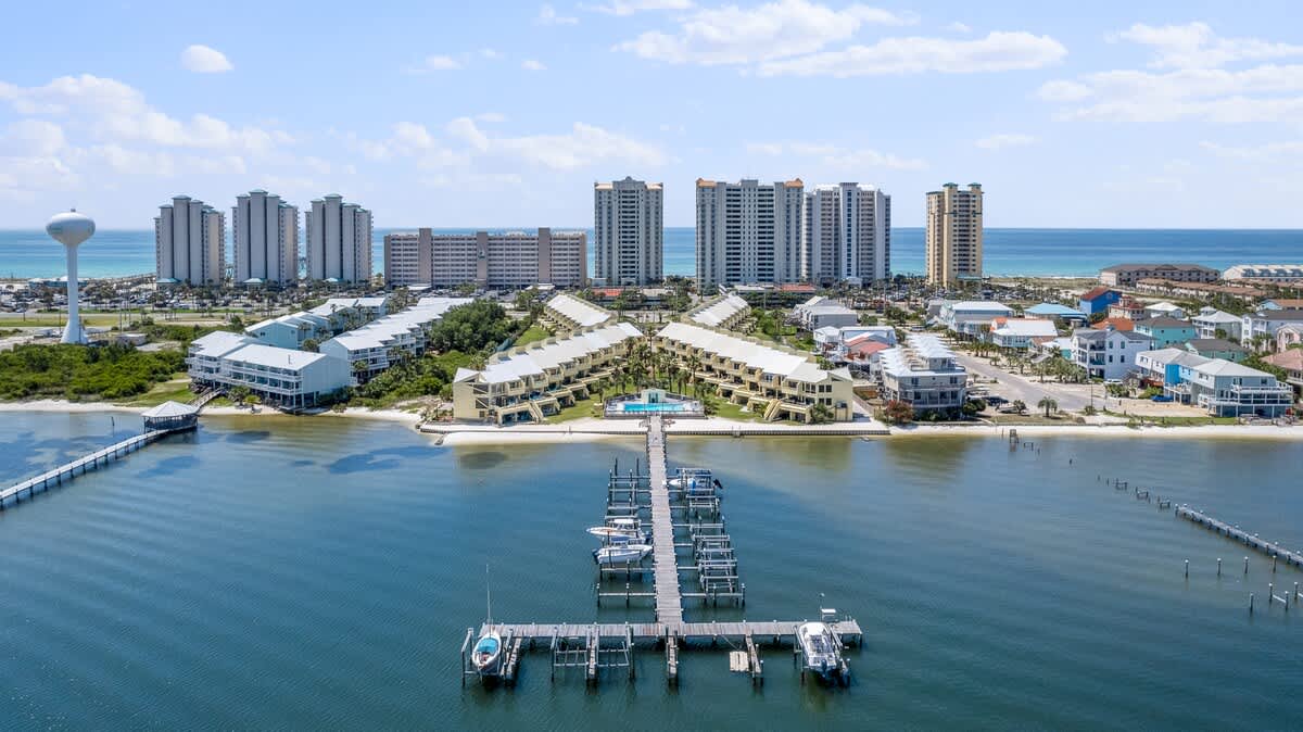 Aerial view of the townhouse complex, highlighting a central swimming pool and a long wooden dock with multiple boat slips extending into the calm blue water. Tall high-rise buildings are visible in the distance across the water.