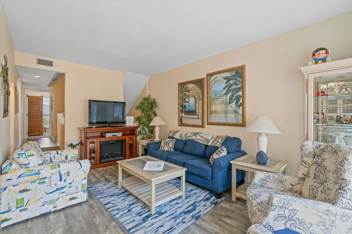 A cozy living room with a blue sofa, a patterned armchair, a TV stand with an electric fireplace, and two large framed artworks on the light beige wall.