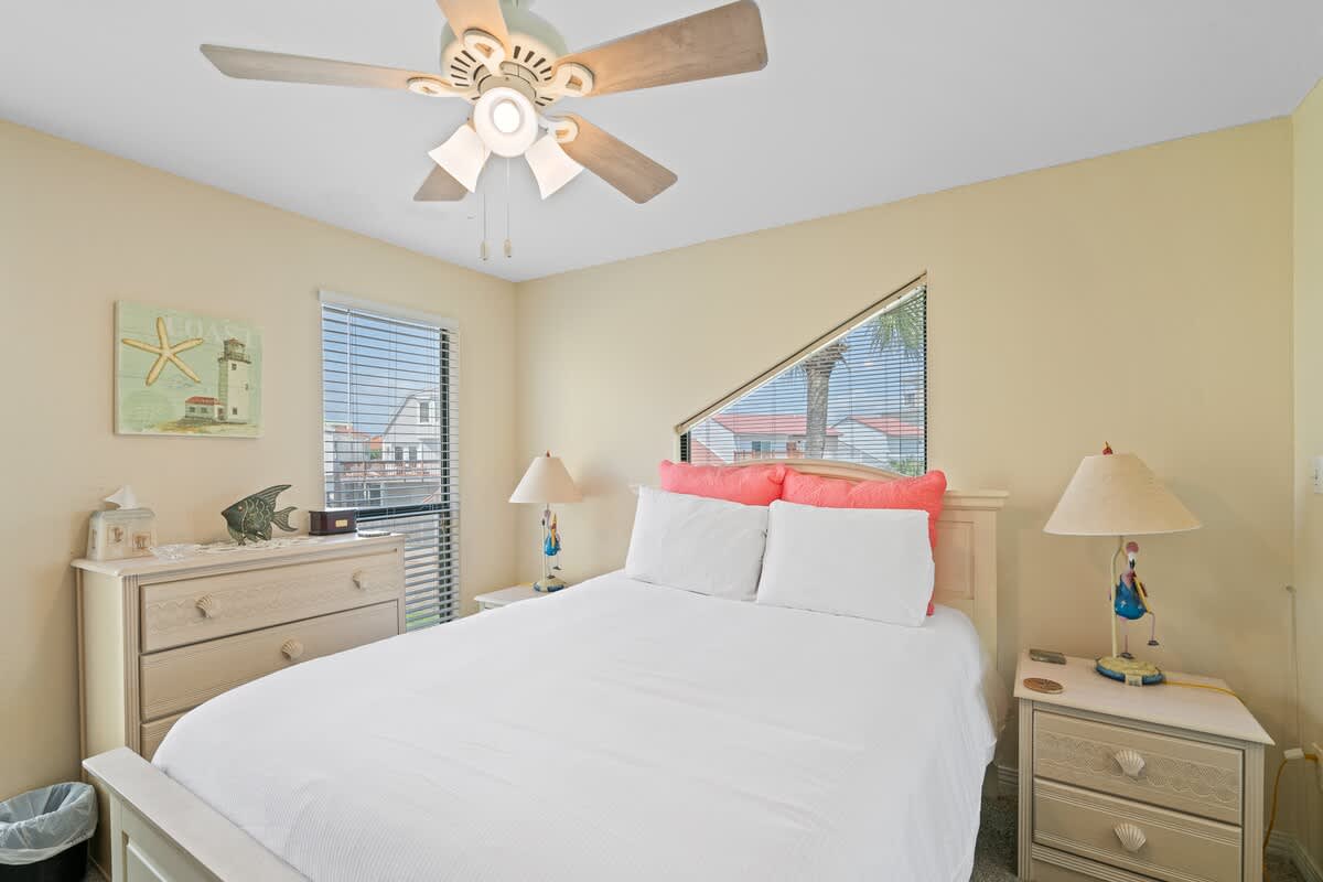 Bright bedroom with a white queen bed, coral pillows, light-colored dresser, and two windows with blinds, featuring a ceiling fan and coastal decor.