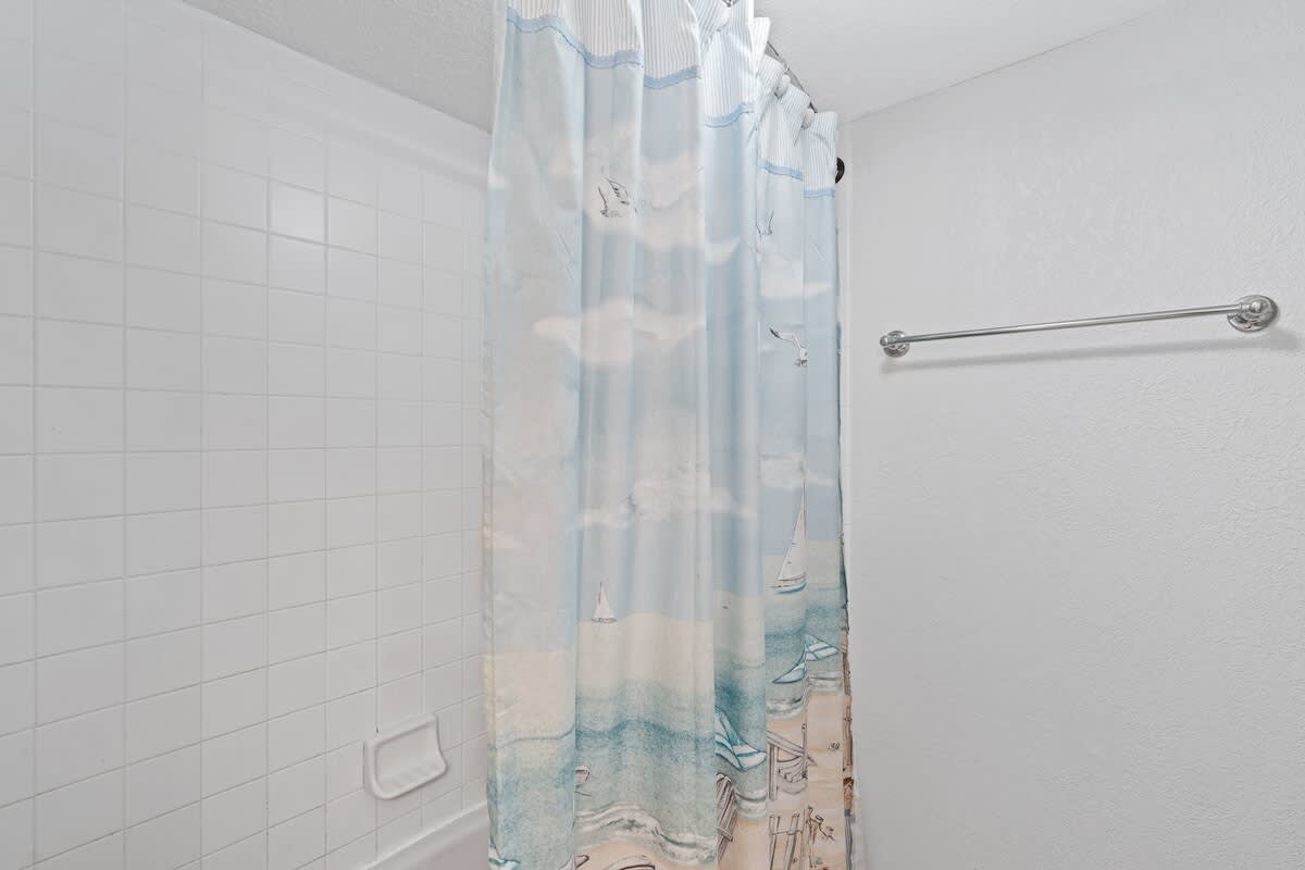 The shower/tub combination in Bathroom-1 is enclosed by white tiled walls and features a vibrant blue and white beach-themed shower curtain, adding a touch of coastal charm.