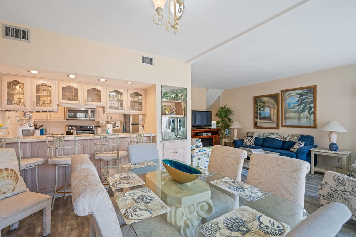 An open-concept living area combines the dining space with a glass table, a kitchen bar with stools, and a cozy living room featuring a blue sofa, framed artwork, and a television.