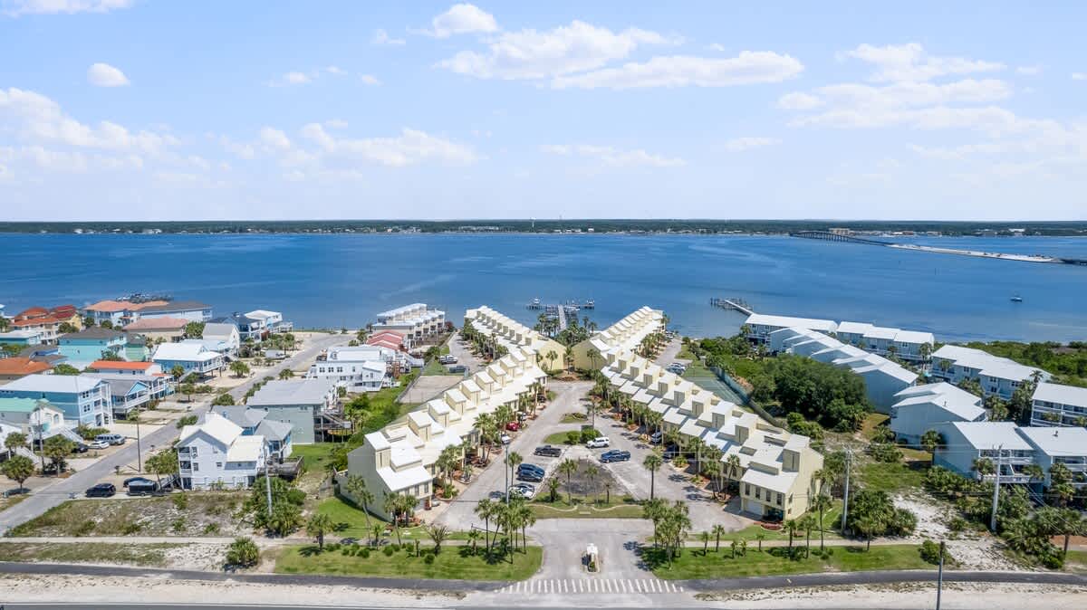 Wide aerial view of a vacation rental complex featuring multiple rows of yellow-sided townhouses with white roofs, arranged in a V-shape. A large body of water with a pier is in the background, and a road runs along the foreground.