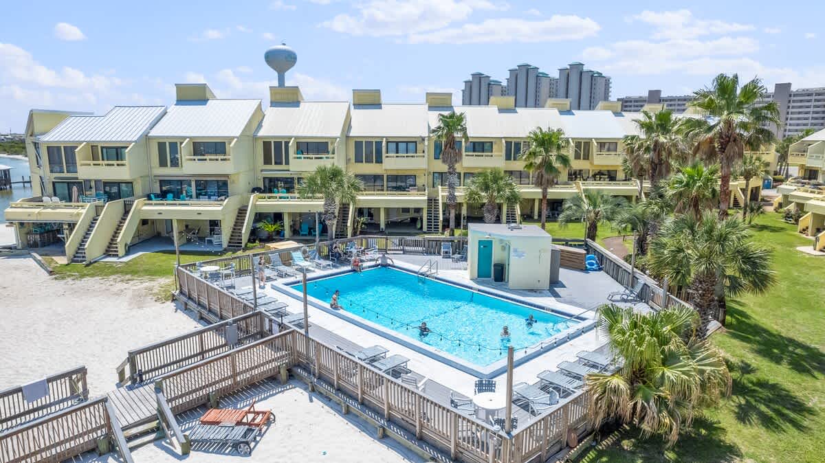 Aerial view capturing the outdoor swimming pool with a surrounding wooden deck and sandy beach area, leading towards the calm bay waters. People are visible in and around the pool, with townhouses in the background.