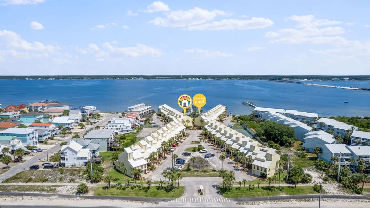 Wide aerial shot of the townhouse complex, revealing its layout with a central drive and the bay stretching into the distance with a bridge. Markers point to the property and the communal pool.