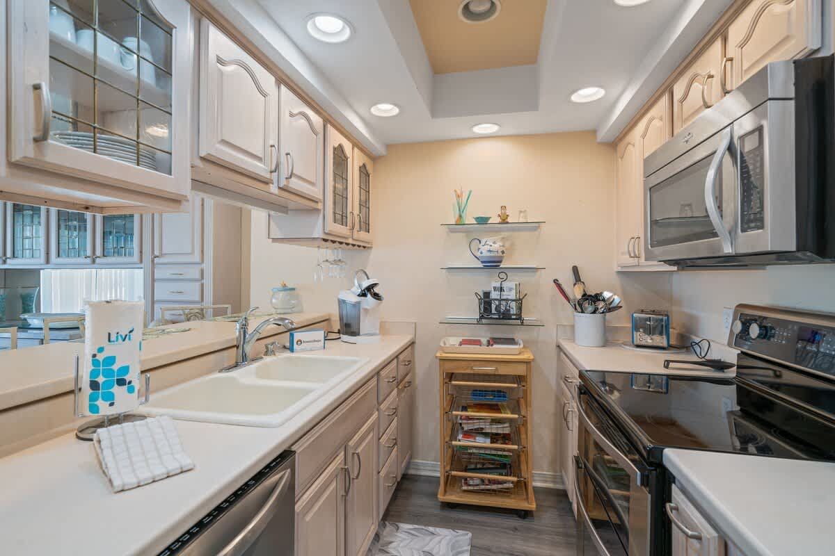 This view of the galley kitchen highlights the double sink and ample white countertop space, with light-colored cabinetry and a mirrored backsplash reflecting the adjacent dining area.