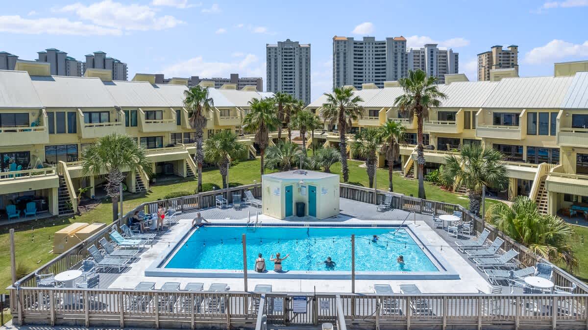 Aerial shot of the vibrant outdoor pool area, featuring a rectangular pool with swimmers and sunbathers on surrounding lounge chairs. Palm trees and the yellow townhouse buildings frame the scene.