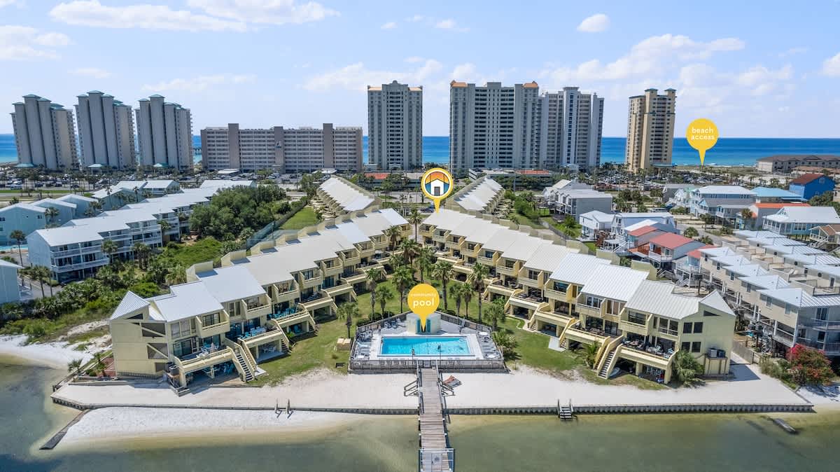 Aerial perspective of the townhouse community, highlighting the central outdoor pool and the adjacent bay, with the ocean and distant high-rise buildings visible. Markers indicate the pool, property, and beach access.