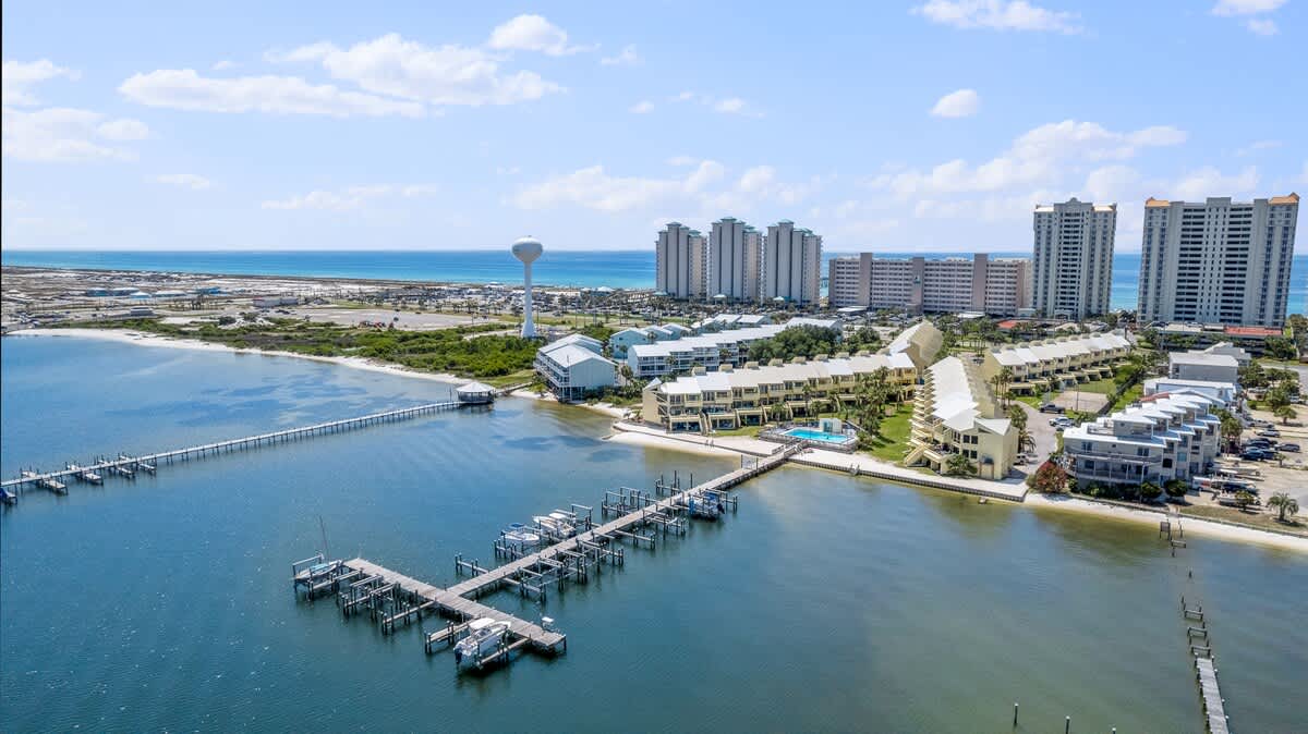 Expansive aerial view of the waterfront townhouse complex, featuring a swimming pool, a long dock with boat slips, and a sandy shoreline. The complex is situated on a bay, with the open ocean and distant high-rise buildings visible beyond a narrow strip of land.