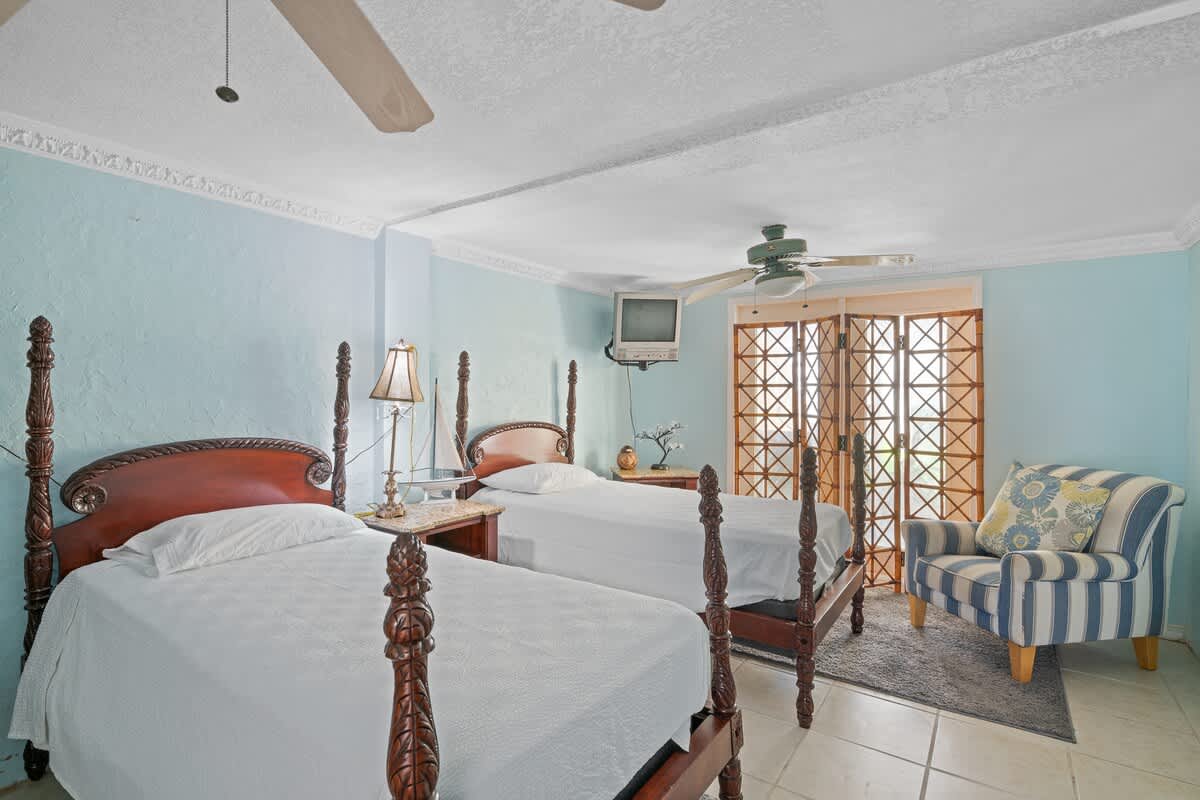 A view of the bedroom showing two dark wood four-poster twin beds with white bedding, light blue walls, tiled flooring, and a striped armchair in the corner next to a folding screen.