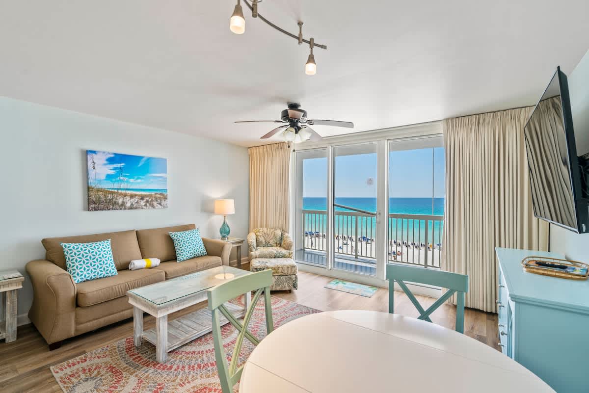 Bright living room with a tan sofa, glass coffee table, and a patterned rug, offering stunning ocean views through floor-to-ceiling glass doors leading to a balcony. A light blue dining table with matching chairs is visible in the foreground.