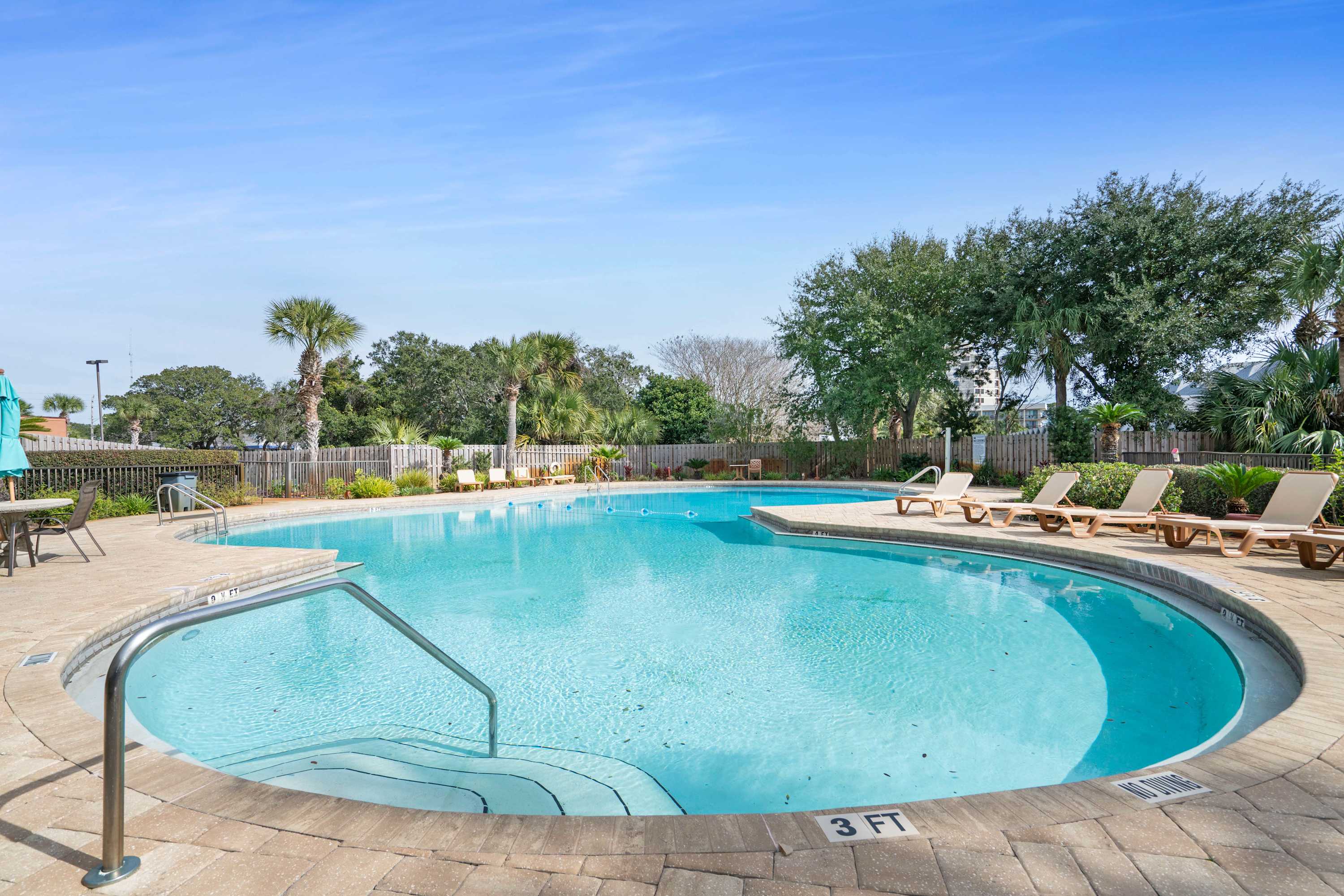 A large, irregularly shaped swimming pool with clear blue water, surrounded by light-colored paver decking and several lounge chairs, set against a backdrop of lush green trees and a bright blue sky.
