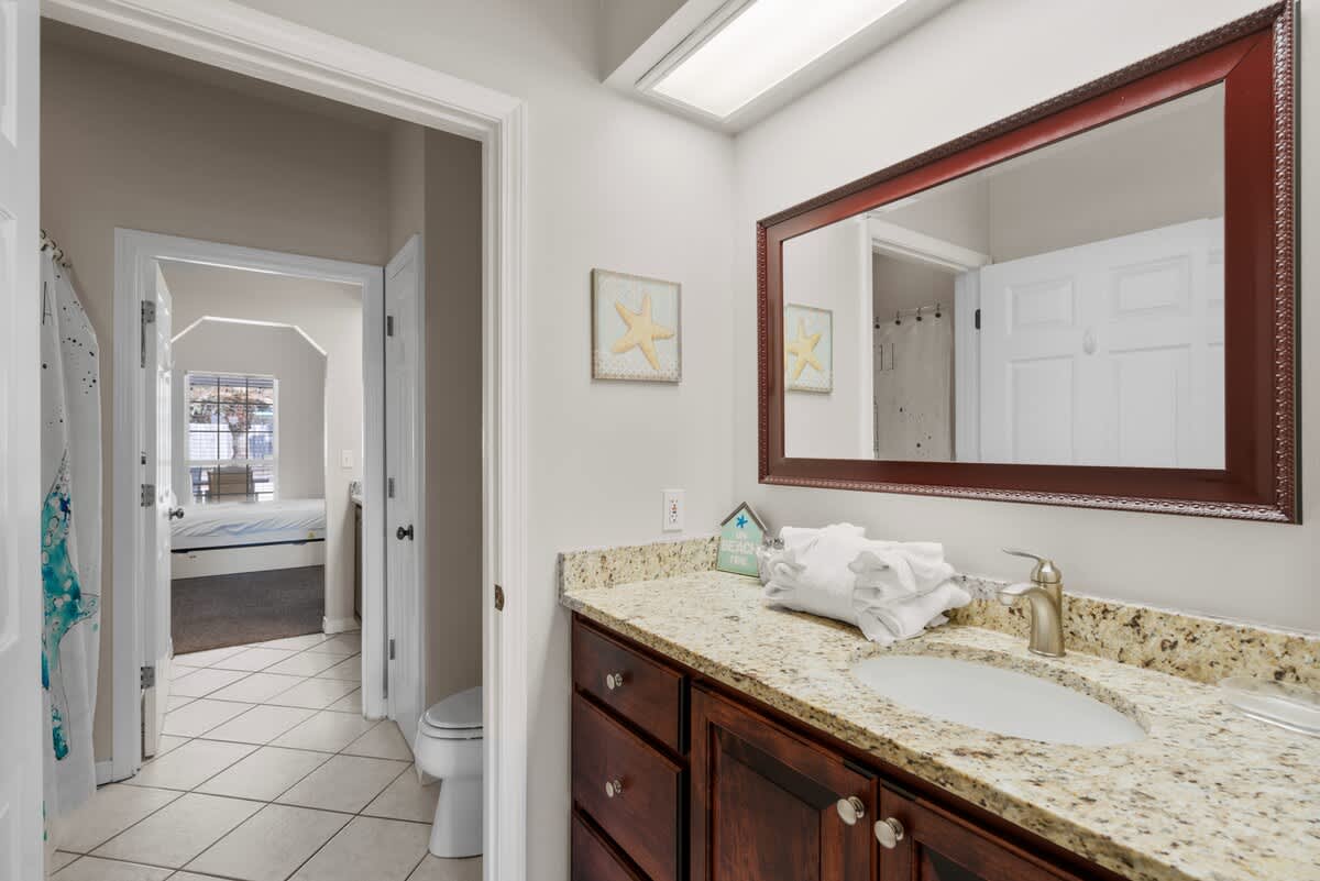 A well-lit bathroom featuring a dark wood vanity with a granite countertop and an undermount sink. A large framed mirror hangs above, and white tiled floors complete the space.