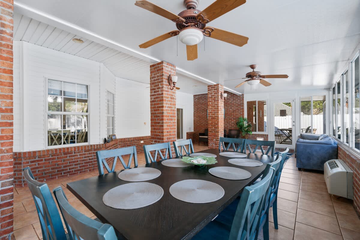 Expansive sunroom featuring a large dining area with a dark table and blue chairs, alongside a cozy seating area. Brick columns and tiled flooring add character, with multiple ceiling fans and bright natural light.