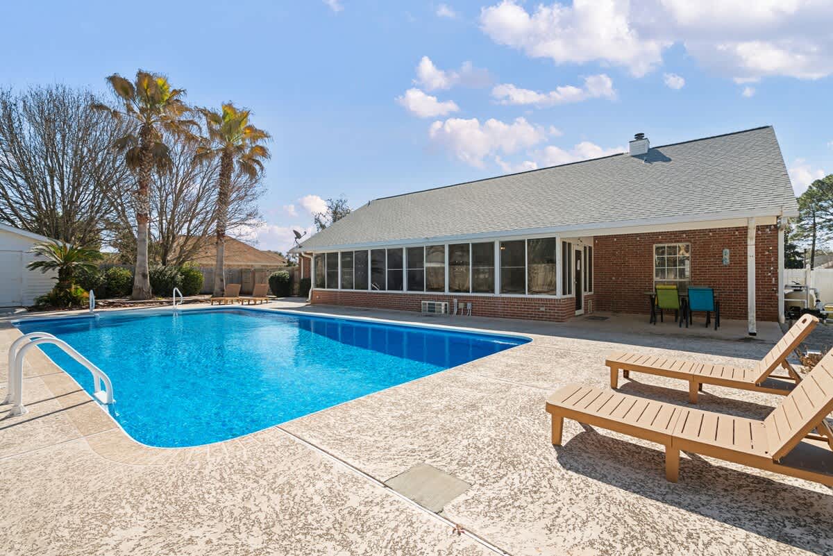 Expansive outdoor swimming pool with bright blue water, surrounded by textured concrete decking and two wooden lounge chairs. A brick house with a large sunroom and palm trees are visible in the background under a clear sky.