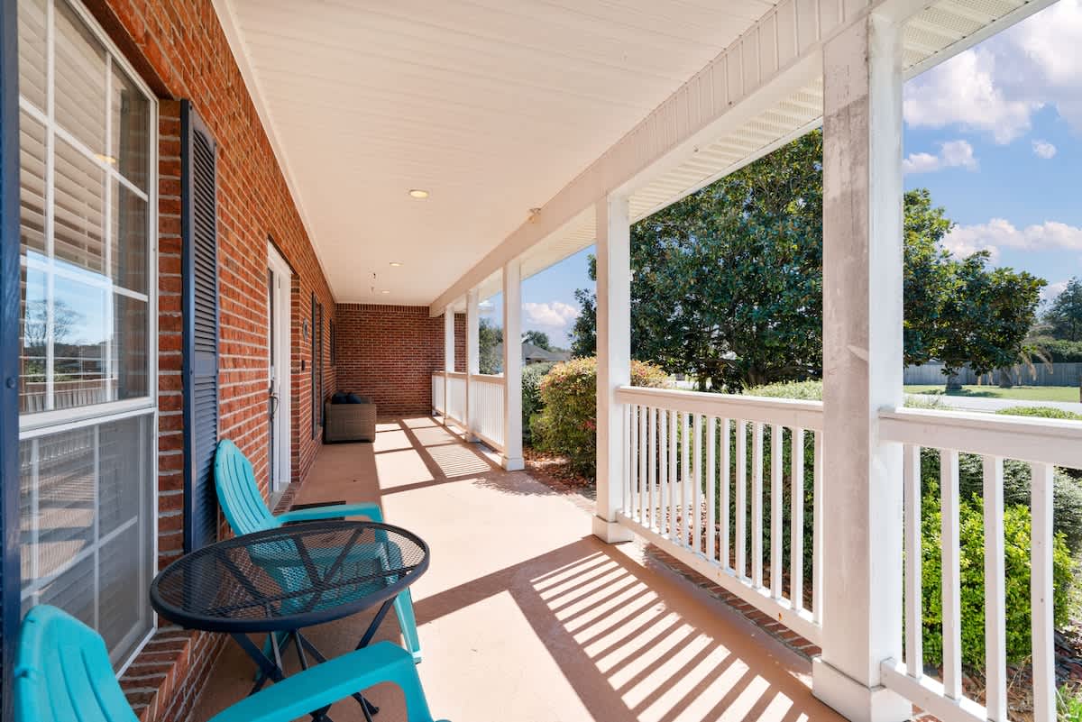 A long, covered front porch with a red brick exterior, white railings, and a small black metal table with two teal chairs, providing a bright and inviting outdoor space.