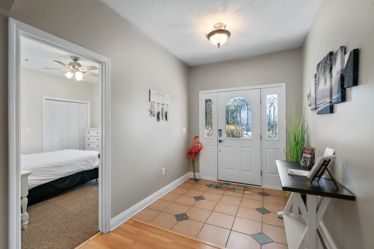 A bright foyer with light gray walls, a white front door featuring decorative glass panels, and a tiled floor with square accents. A small console table with artwork and a red flamingo statue add decorative touches.