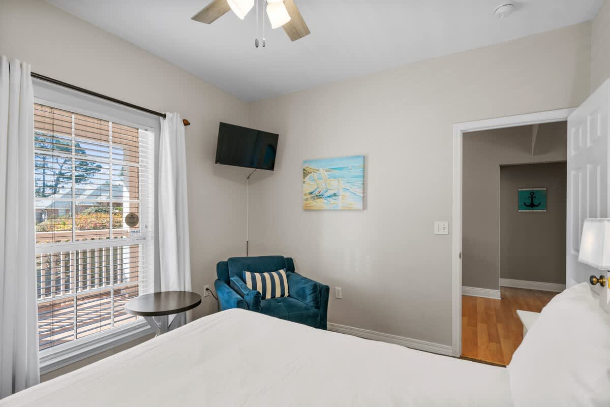 A cozy bedroom featuring a white-covered bed, a wall-mounted TV, and a blue armchair next to a round side table. A large window with blinds and white curtains overlooks an outdoor deck.