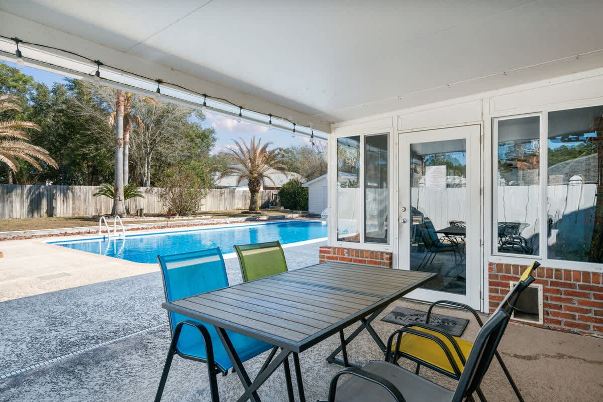 Covered outdoor patio area featuring a dark gray dining table with colorful chairs, overlooking the bright blue swimming pool. Palm trees and a white fence are visible in the sunny backyard.