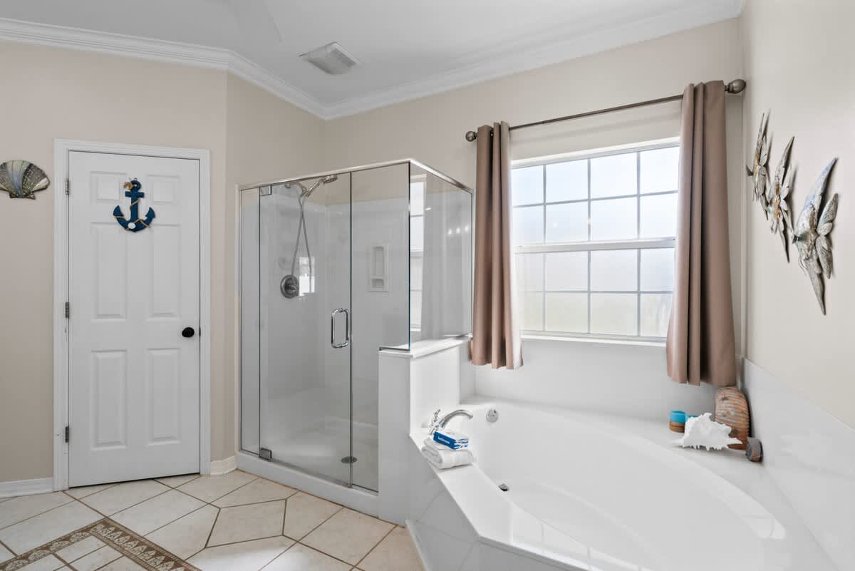 A bright corner of the master bathroom, showcasing the glass-enclosed walk-in shower and a large corner bathtub positioned under a window with blinds, adorned with nautical wall decor.