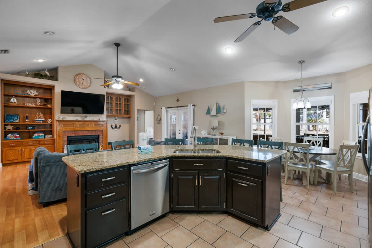 View of the kitchen island with granite countertops and dark cabinets, looking towards a dining area with a round table and chairs. The living room with a blue sectional is visible in the background.