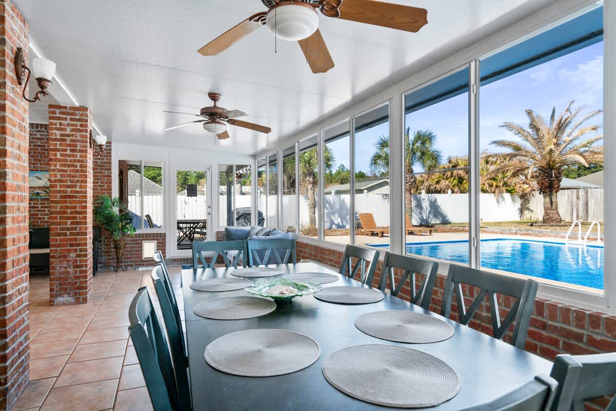 Spacious sunroom with a long dark dining table and blue chairs, set on terracotta-colored tiled flooring. Large windows provide clear views of the outdoor pool and tropical landscaping, with a ceiling fan overhead.
