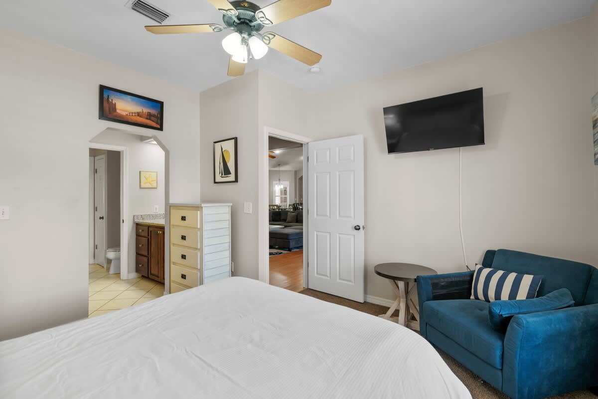 A comfortable bedroom with a white-covered bed, a blue armchair, and a wall-mounted TV. A light-colored dresser and a doorway leading to an adjacent bathroom are visible.